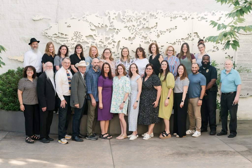 A diverse group of about 25 adults stands closely together outdoors, smiling at the camera. They are posing in front of a decorative wall with a map and Hebrew text. Trees and plants frame the scene.