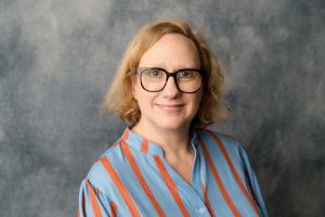 A woman with shoulder-length blonde hair, wearing black glasses and a blue shirt with orange vertical stripes, stands in front of a gray mottled background, smiling gently at the camera.