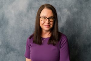 A woman with long straight brown hair, wearing glasses, a purple dress, and a necklace, smiles softly while standing in front of a gray textured background.